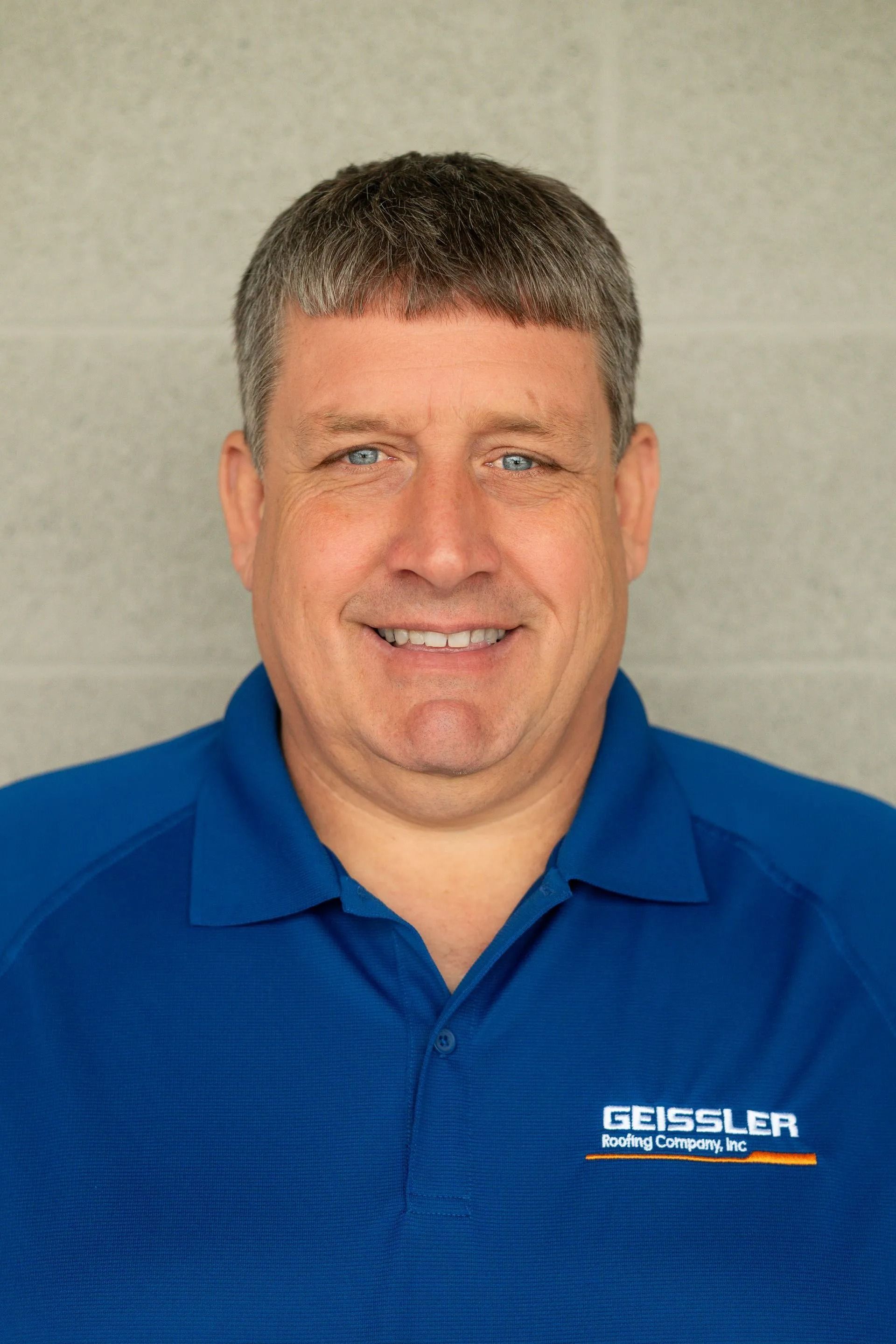 A person with short brown hair smiling while wearing a blue collared shirt with a company logo against a gray wall.