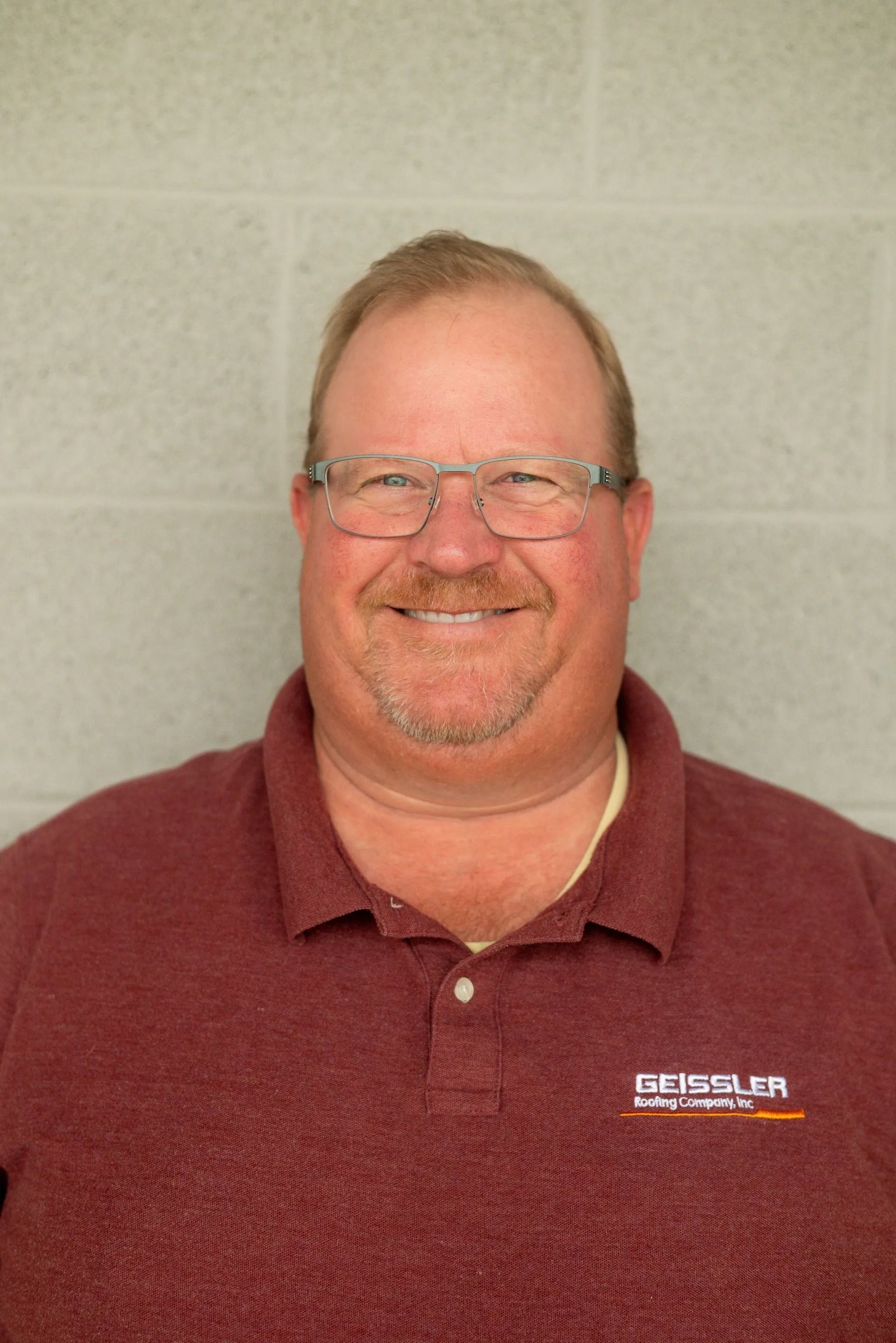 A person wearing glasses and a maroon polo shirt with a logo, smiling in front of a gray cinder block wall.