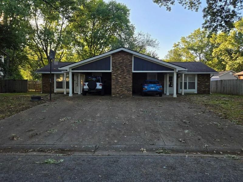 A single-story duplex with a shared stone chimney, two covered carports, and a large concrete driveway under trees.