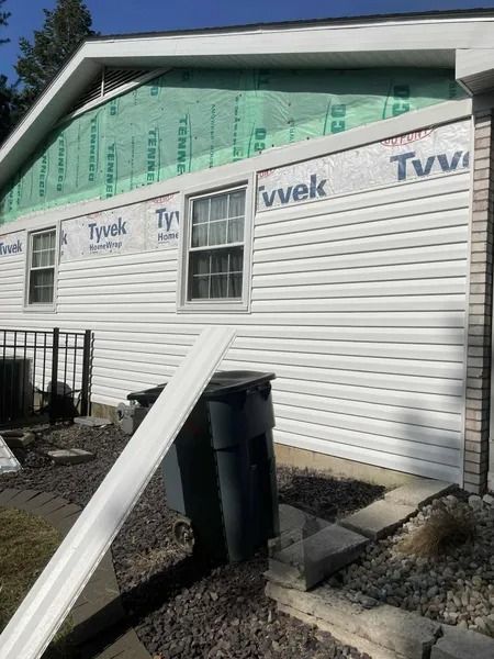A side view of a house undergoing siding installation, with white vinyl siding, exposed Tyvek wrap, and a piece of trim.