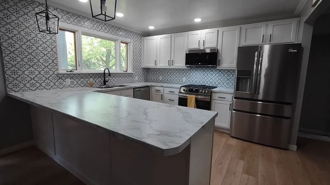 A modern kitchen with white cabinets, marble countertops, stainless steel appliances, and a patterned tile backsplash.