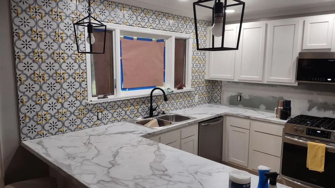 A kitchen with white cabinets, marble-patterned countertops, and patterned tiled walls around a window and sink.