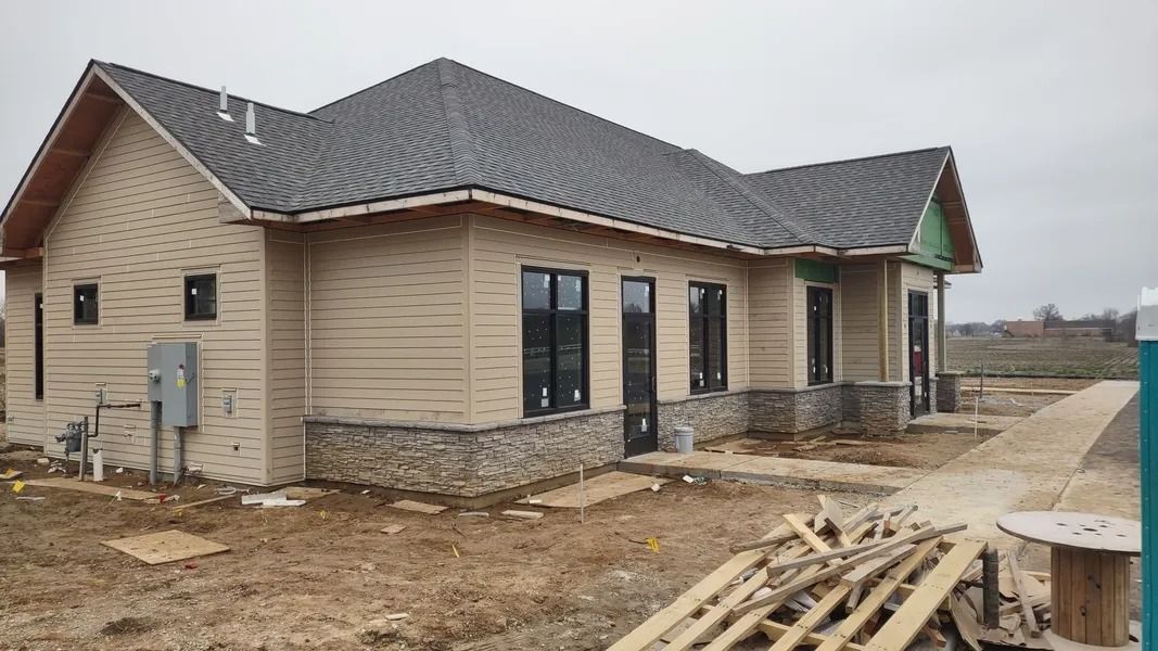 A side view of a new tan-sided house under construction with stone accents, dark windows, and a gray shingled roof.