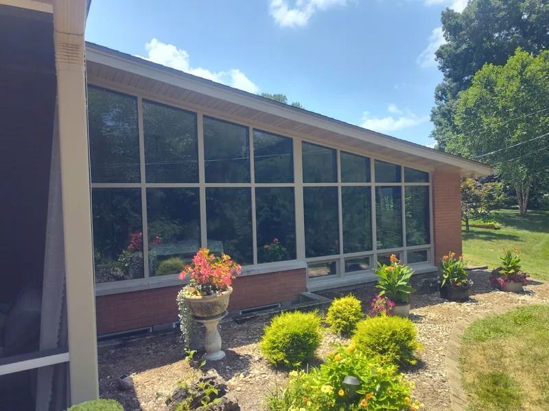 A house exterior featuring a large, slanted glass window wall above a brick foundation, set beside a sunny grassy lawn.