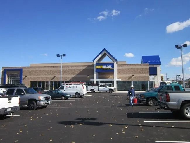 A tan brick CarMax building under a clear blue sky, with a parking lot containing several vehicles and a person walking.