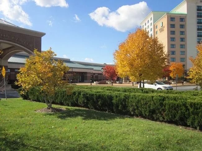 A hotel building with a tan facade and a tall tower stands behind a green lawn, trimmed hedges, and autumn-colored trees.