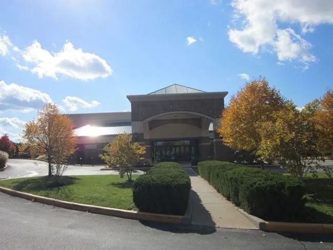 A modern commercial building with a tan facade and a central entrance, framed by yellow autumn trees and green shrubs.