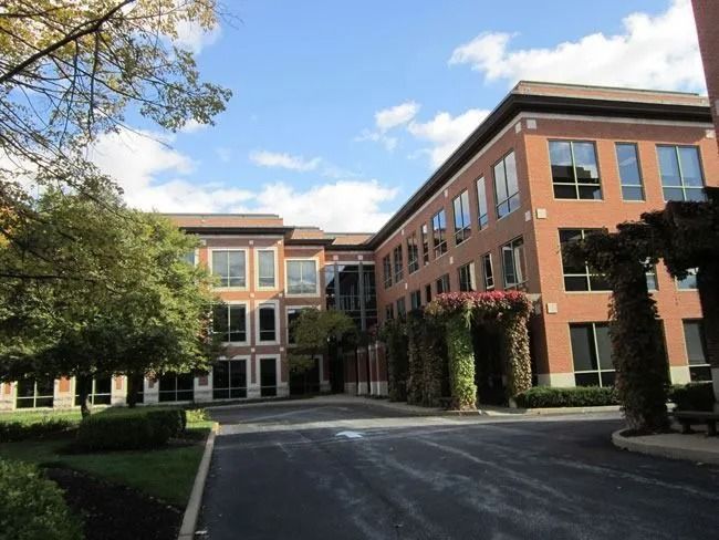 A multi-story red brick office building with a paved driveway and landscaped trees under a sunny blue sky.
