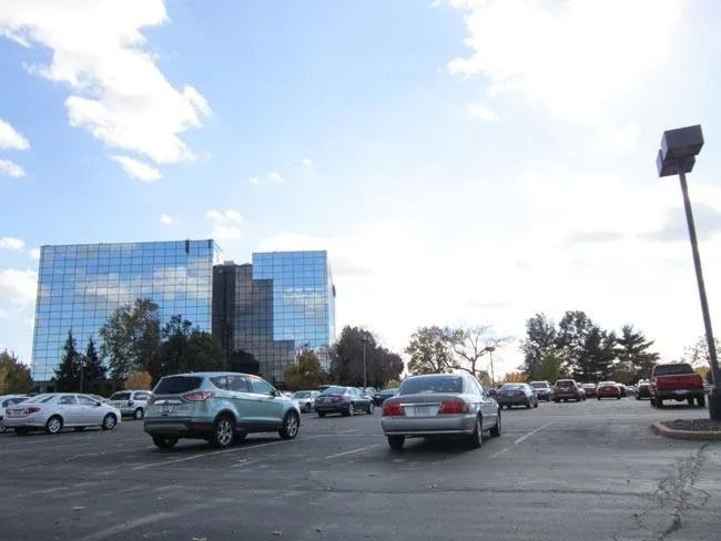 Cars parked in a paved lot in front of a modern, multi-story glass office building under a blue, cloudy sky.