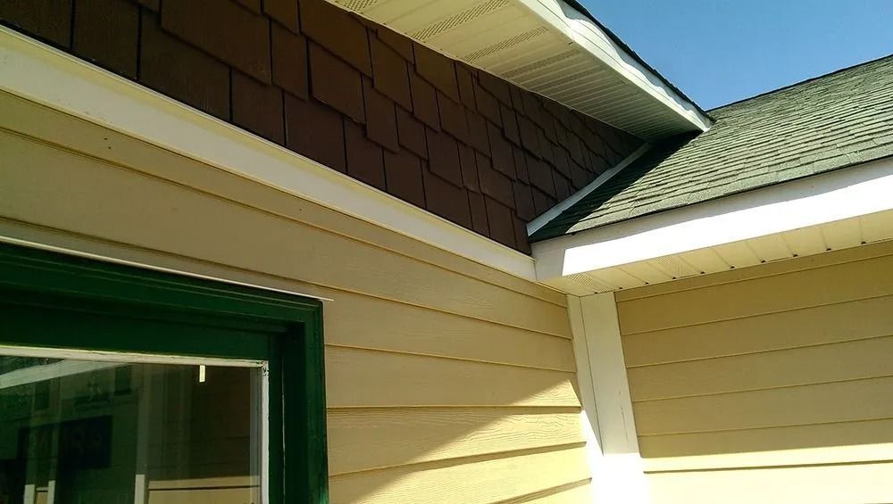 A home exterior showing a corner junction with brown shingle siding, beige lap siding, white trim, and a green window.