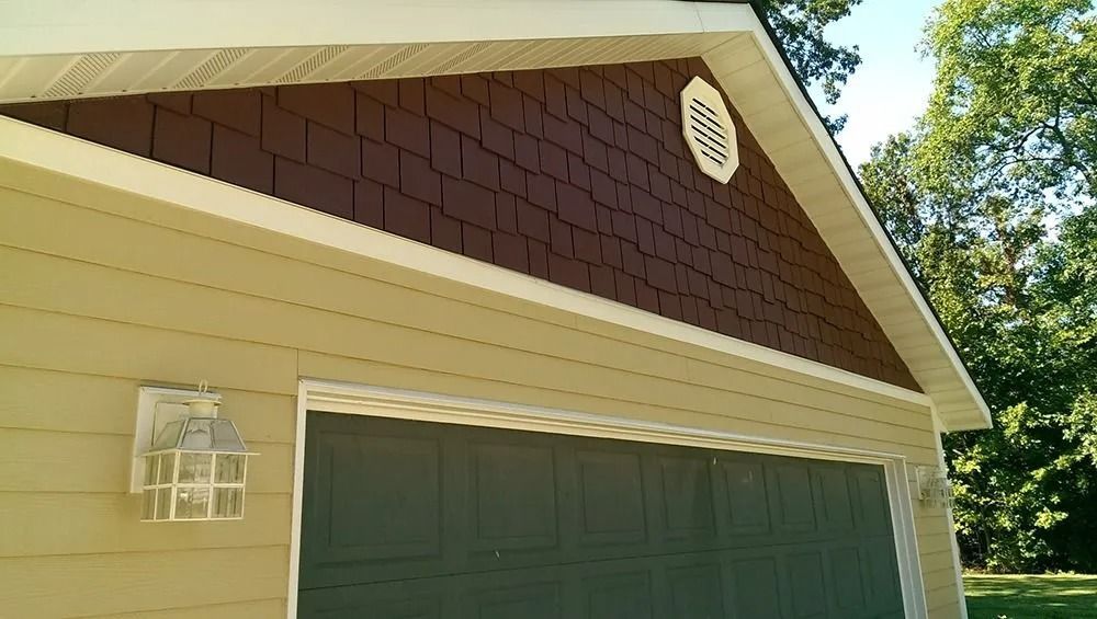 A beige house exterior with a garage, dark green garage door, and brown shake siding on the triangular upper gable.