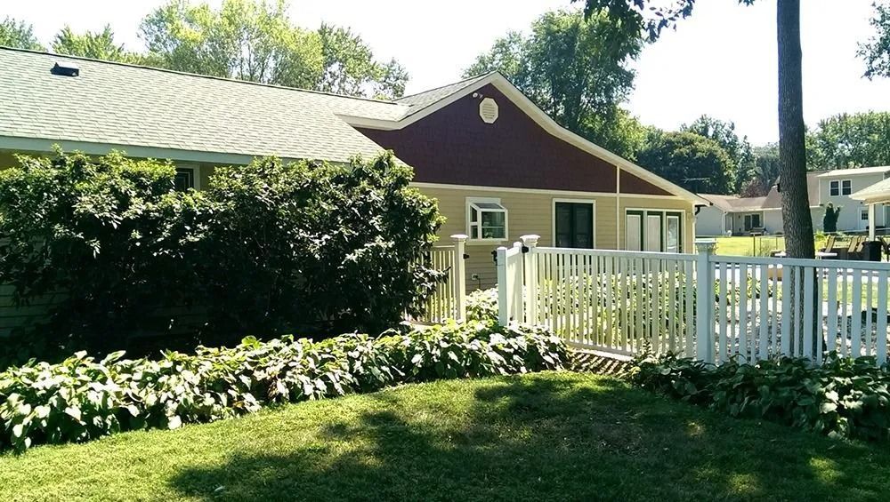 A single-story house with tan and dark red siding behind a white fence, surrounded by lush green bushes and trees.