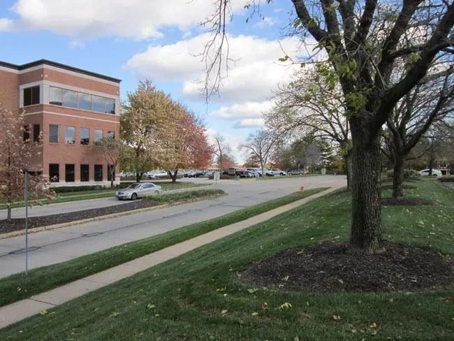 A multi-story brick office building sits beside a paved parking lot and a lawn with trees under a partly cloudy sky.