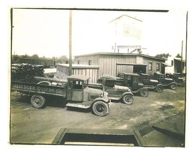 A fleet of vintage trucks parked in front of a building with a large water tower, likely a industrial or warehouse site.
