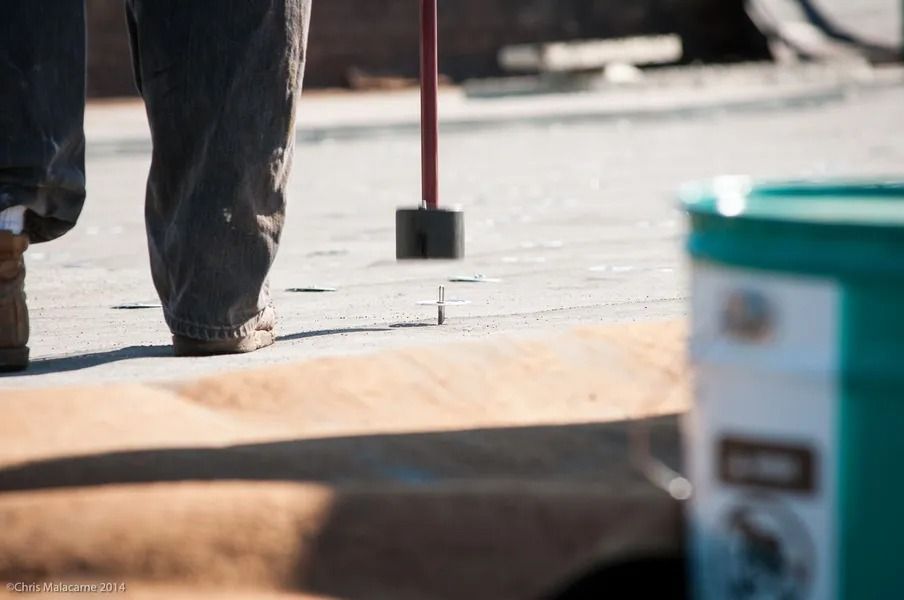 A construction worker uses a magnetic tool to collect nails on a flat, paved surface next to a teal bucket.
