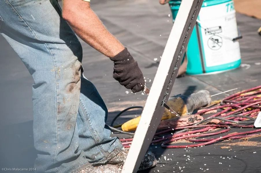 A construction worker in jeans and a black glove cuts a long, white board, scattering debris on a dark rooftop.