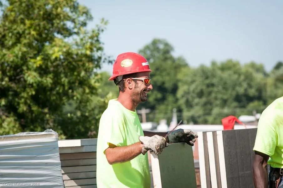 A construction worker in a red hard hat and neon yellow shirt smiles while holding a white construction material panel.