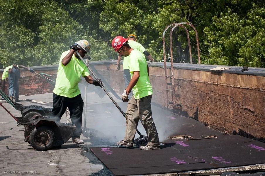 Two workers in bright neon shirts and safety helmets use long tools to spread hot roofing material on a sunny rooftop.