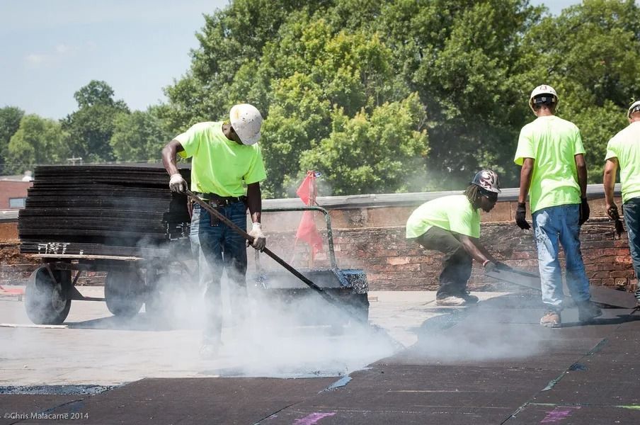 Construction workers in neon shirts apply hot roofing material, creating steam on a sunny rooftop.