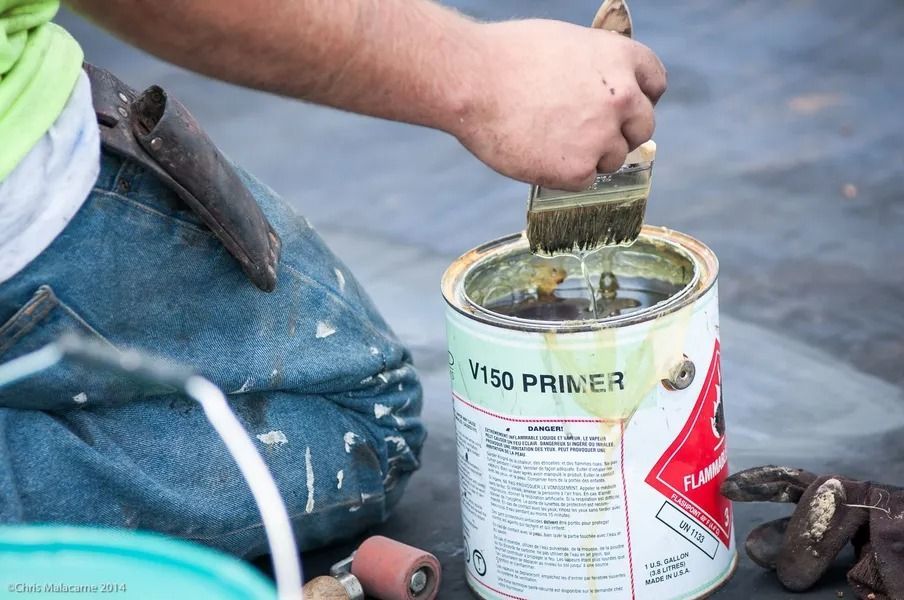 A worker in blue jeans dips a paintbrush into a metal can labeled V150 Primer on a rooftop.