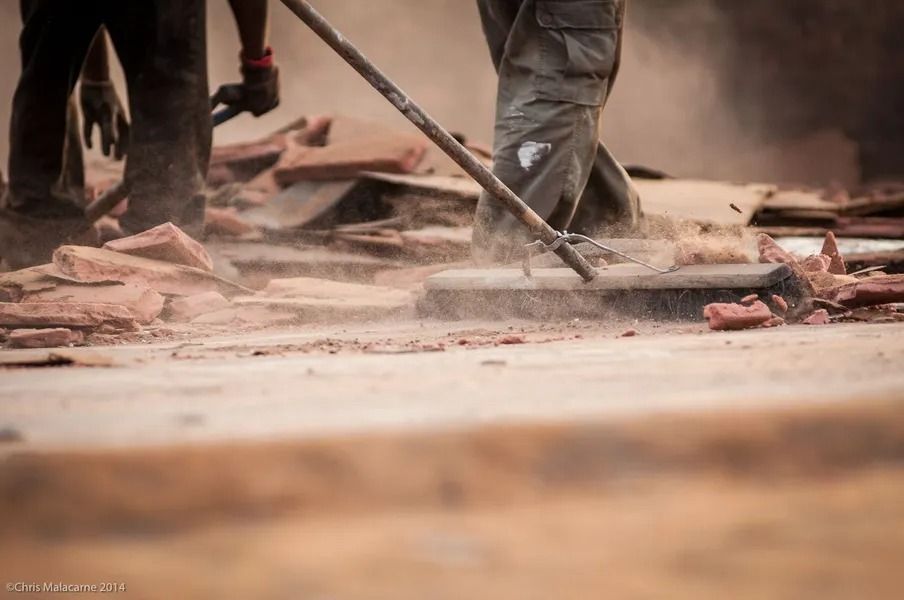 Workers clearing dust and debris from the ground with long-handled tools in a construction setting.