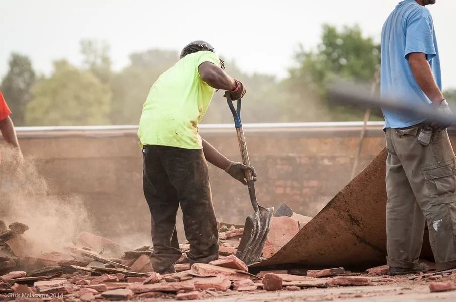 Two construction workers clear debris and red roof tiles from a building site.