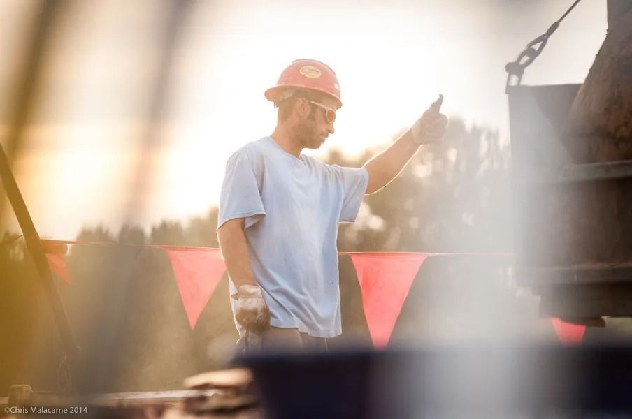 A worker in a hard hat and light-colored shirt gives a thumbs up on a construction site at sunset.