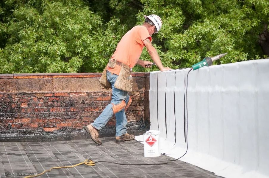 A worker in a hard hat uses a heat tool to seal white roofing material against a brick wall on a flat roof.