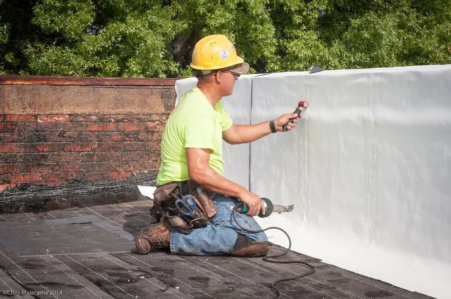 A worker in a yellow hard hat and neon shirt kneels on a rooftop using a hand-held heat welder on white roofing material.