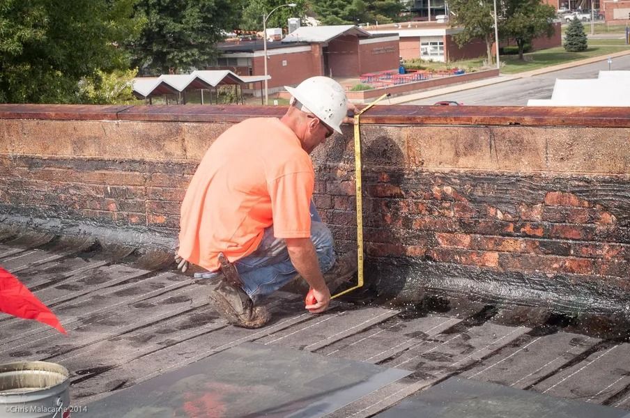 A construction worker in a white hard hat and orange shirt kneels on a flat roof, measuring a brick wall with a tape.