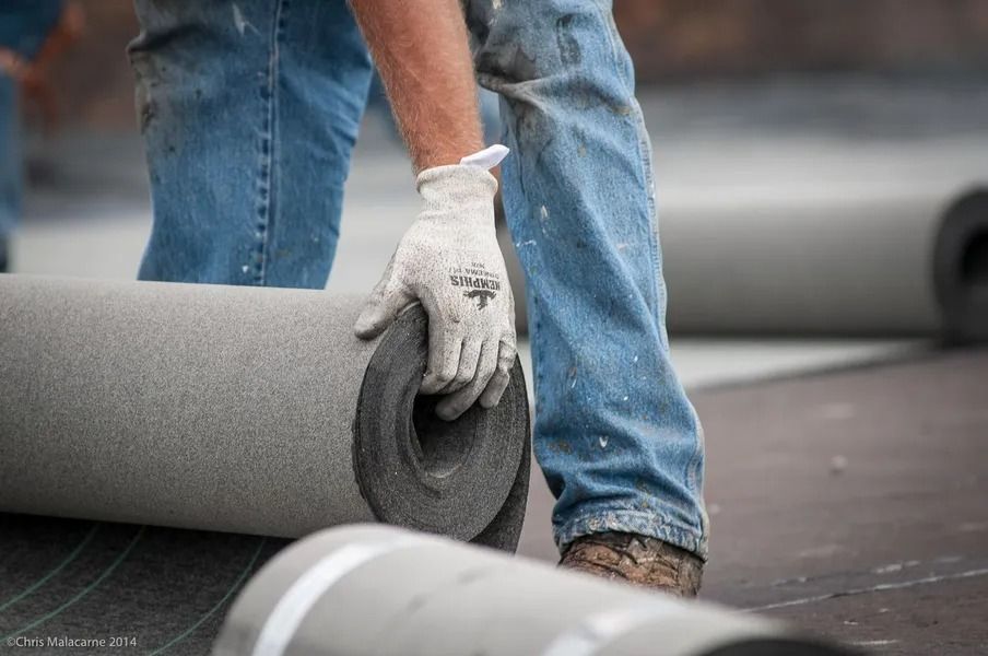 A worker in jeans and work gloves unrolls a textured gray roofing material on a flat surface.