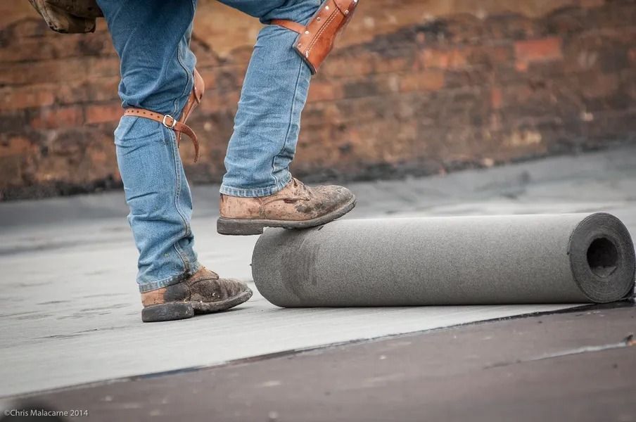 A worker wearing knee pads stands on a rooftop, one foot resting on a rolled layer of gray roofing material.