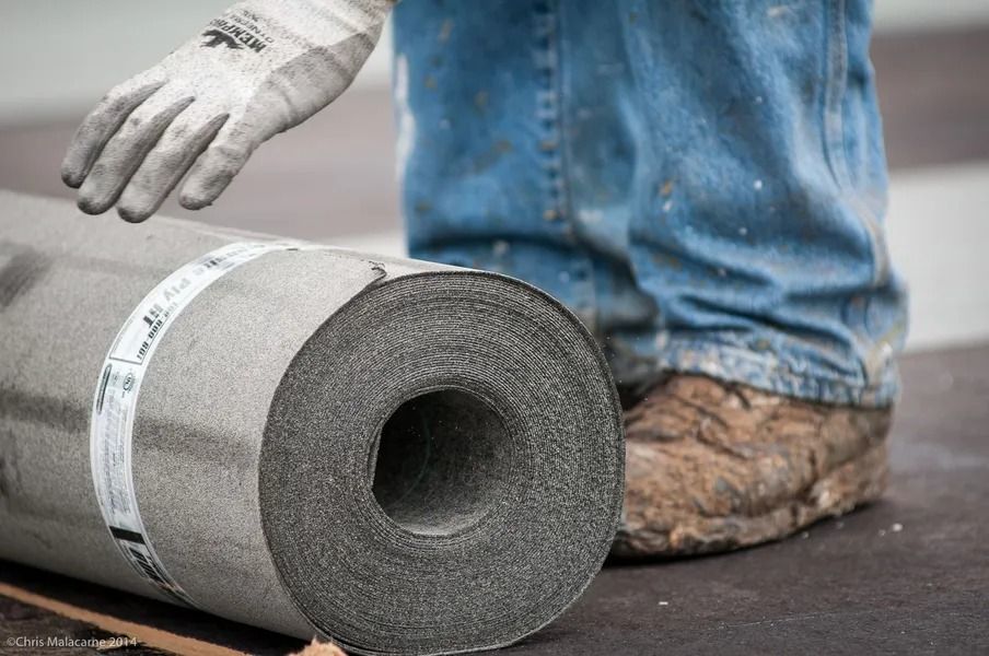 A close-up of a worker in jeans and work boots placing a hand on a roll of dark, textured roofing material.