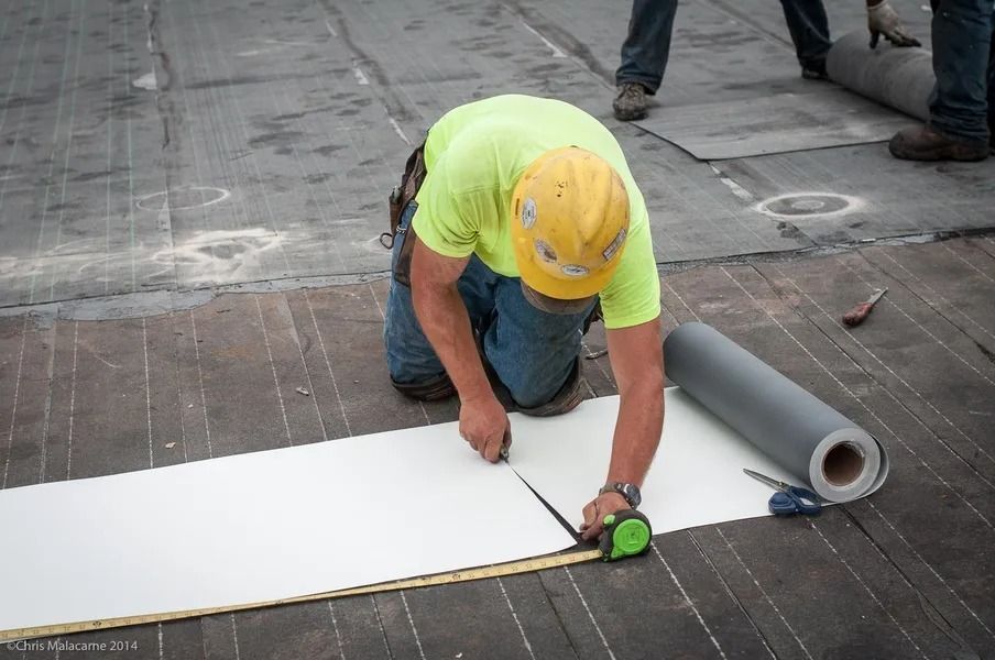 A worker in a neon yellow shirt and hard hat cuts a roll of white roofing material on a dark surface.