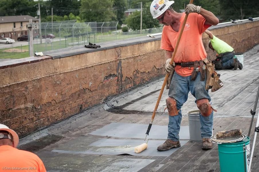 Workers in high-visibility orange and yellow shirts applying sealant with a roller on a flat commercial rooftop.