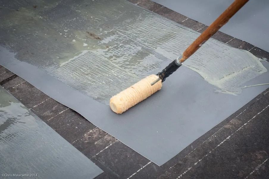 A paint roller spreads a light gray adhesive onto a dark, flat roofing membrane.