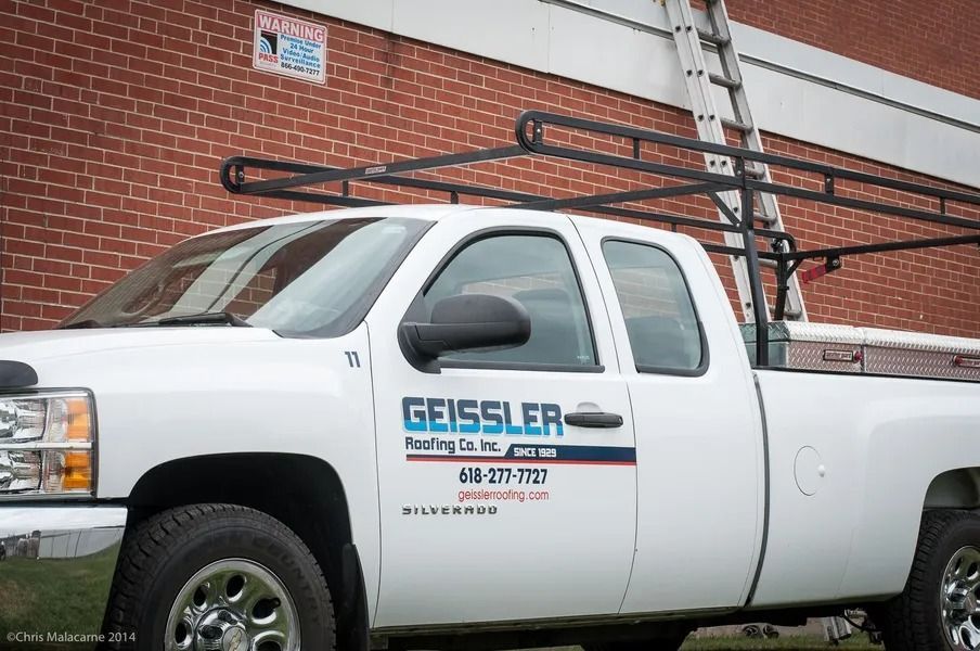 A white Geissler Roofing work truck with a ladder rack parked in front of a red brick building.