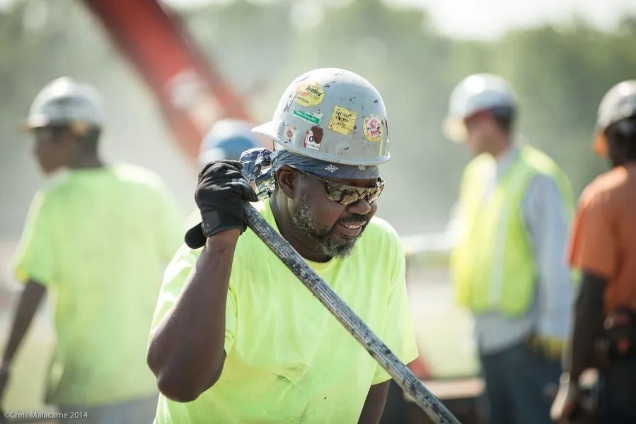 A construction worker in a high-visibility yellow shirt and hard hat carries a tool over their shoulder at a job site.