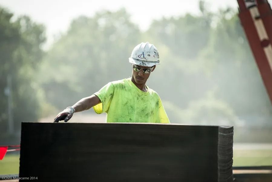Construction worker in a high-visibility yellow shirt and hard hat gesturing toward a stack of black panels outdoors.