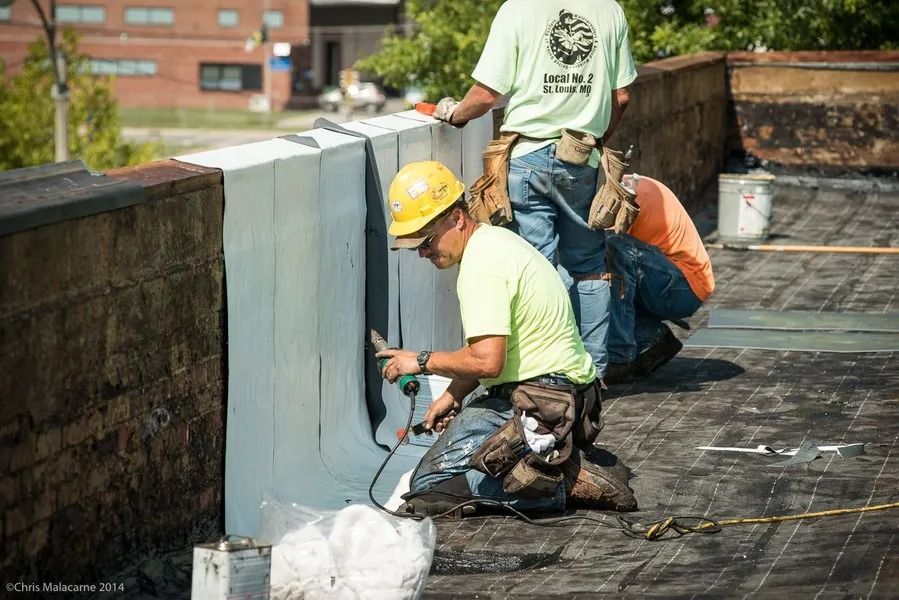 Construction workers in bright shirts and helmets installing waterproof roofing materials along a brick wall on a rooftop.