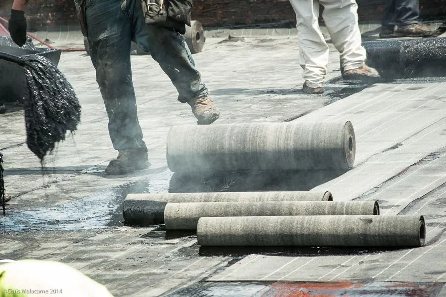 Construction workers apply black roofing felt rolls to a flat roof with hot tar.