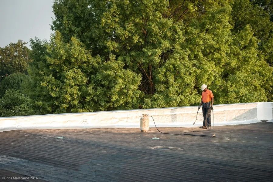 A worker in an orange shirt and hard hat uses a torch to apply roofing material on a flat roof near trees.
