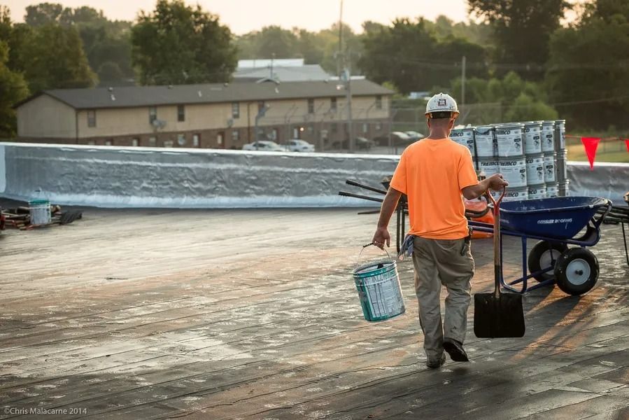 A worker in an orange shirt and hard hat walks across a rooftop, carrying a bucket and shovel near a construction cart.