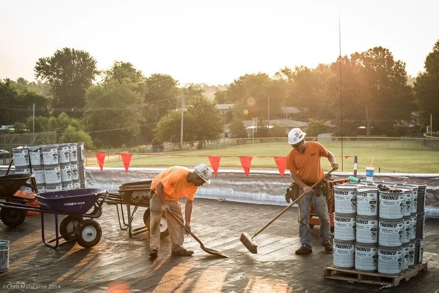 Two construction workers in orange shirts work on a flat roof, one sweeping and the other using a shovel near supplies.