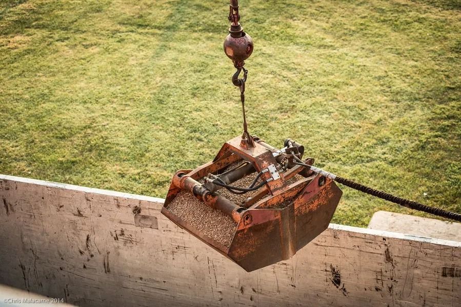 A rusty metal orange crane grab bucket hangs suspended over a light-colored concrete barrier with green grass in the back.