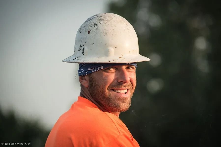 A smiling person wearing a hard hat and an orange high-visibility shirt, seen from the side against a blurred background.