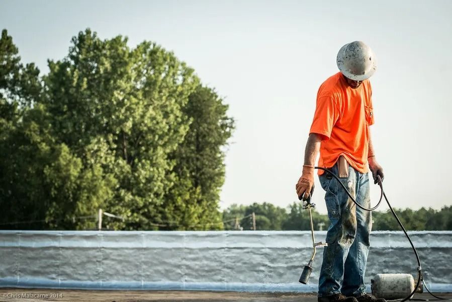 A worker in an orange shirt and hard hat holds a roofing torch on a rooftop with trees in the background.