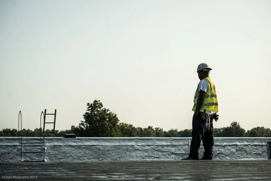 A construction worker in a high-visibility vest and hard hat stands on a flat roof next to a metal ladder.