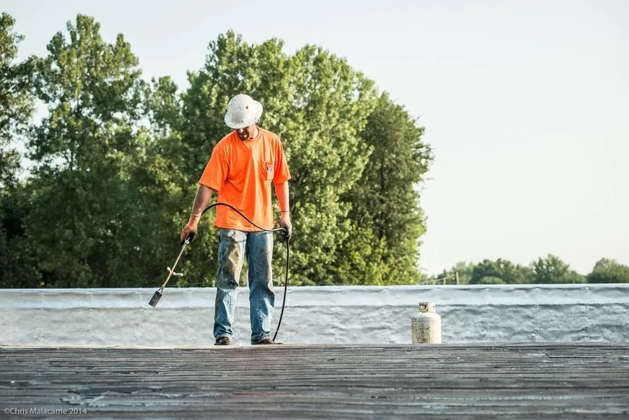 A worker in an orange shirt and hard hat uses a propane torch to heat a flat roofing surface.