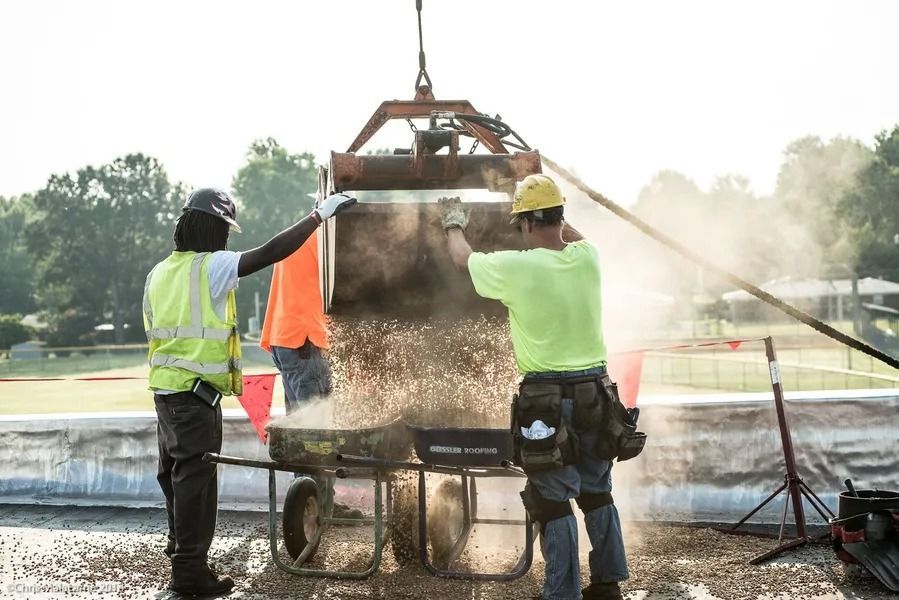 Three workers in high-visibility safety gear guide a crane-suspended bucket pouring gravel into a wheelbarrow on-site.